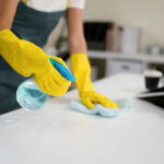 Young asian woman wearing yellow gloves and an apron, smiling while cleaning a white table with disinfectant spray and a blue cloth, promoting hygiene and cleanliness in her workspace.
